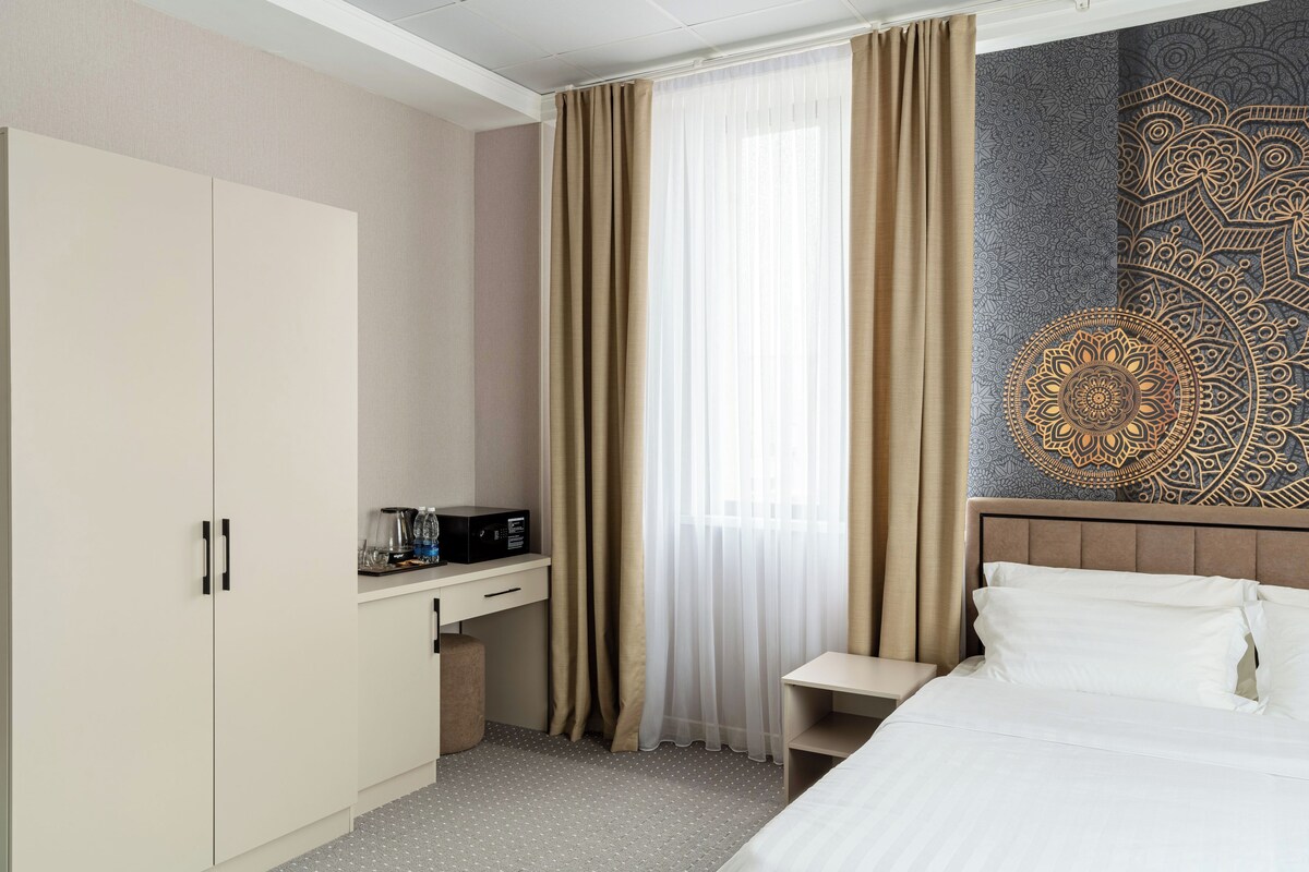 A hotel room with a bed, a desk with a coffee maker, a wardrobe, and a patterned wall.