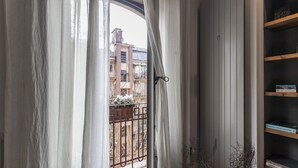 Vista a través de una ventana con cortinas ligeras, que muestra un balcón con barandilla y un edificio al fondo.