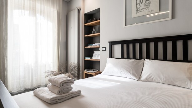Una cama bien hecha con sábanas blancas, una mesita de noche con lámpara y una estantería con diversos objetos.