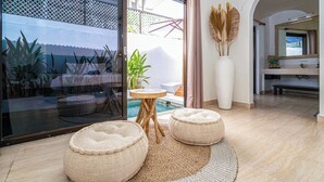 A modern living room with a glass door leading to a pool area, a round wooden table, two round ottomans, and a large vase with dried plants.