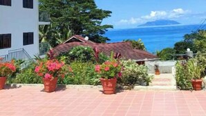 Un bâtiment blanc doté de nombreuses fenêtres et d’un balcon, entouré de plantes en pot et offrant une vue sur l’océan.