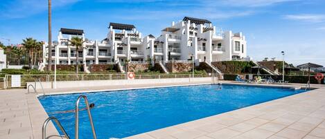 A swimming pool with a clear blue water, surrounded by a tiled deck, and a building with multiple balconies in the background.