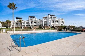 Ein Swimmingpool mit klarem, blauem Wasser, umgeben von einer gefliesten Terrasse, und ein Gebäude mit mehreren Balkonen im Hintergrund.