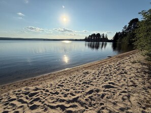 A sandy beach with calm water, trees, and a clear sky.