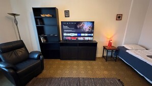 Ein Zimmer mit einem schwarzen Ledersofa, einem an der Wand befestigten Fernseher, einem kleinen Tisch mit einer Lampe und einem Bett mit weißer Bettwäsche.