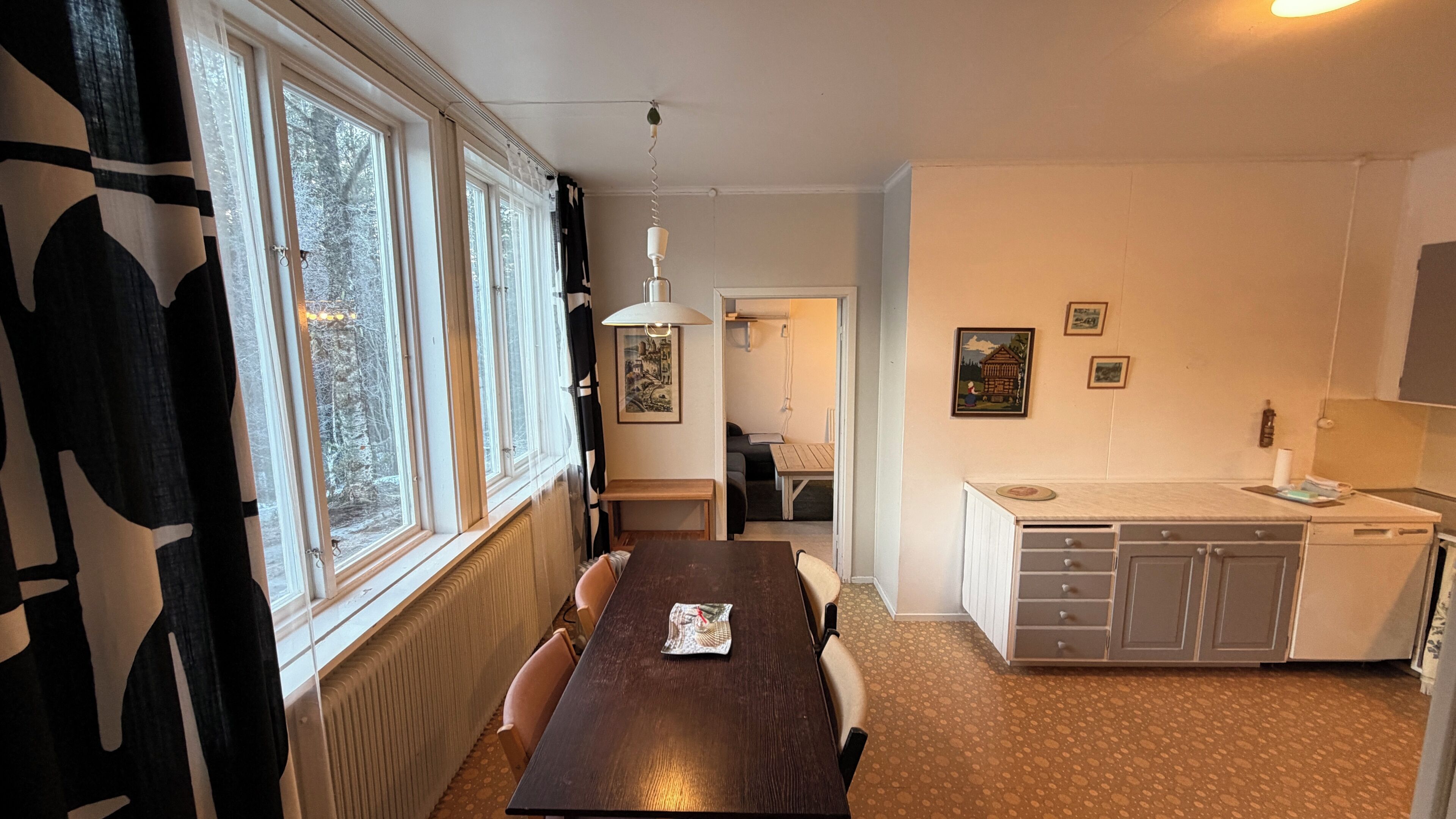 Uma cozinha com mesa de jantar, cadeiras, radiador, janela com cortinas e um painel com quadros.
