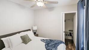 Una habitación de hotel con cama, ventilador de techo, mesita de noche con lámpara y baño visible a través de una puerta abierta.