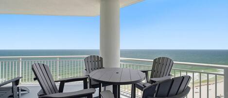 Un balcon avec une table ronde et quatre chaises Adirondack, donnant sur une plage et l’océan.
