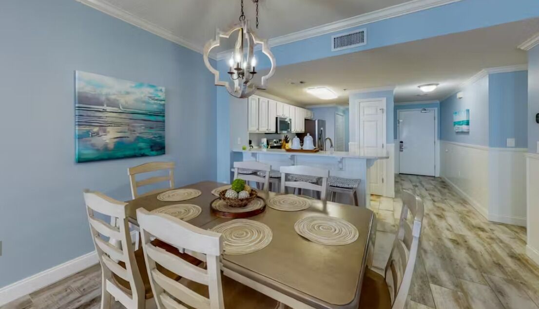 A dining area with a round table, chairs, and a chandelier. There is a kitchen in the background with white cabinets and appliances.