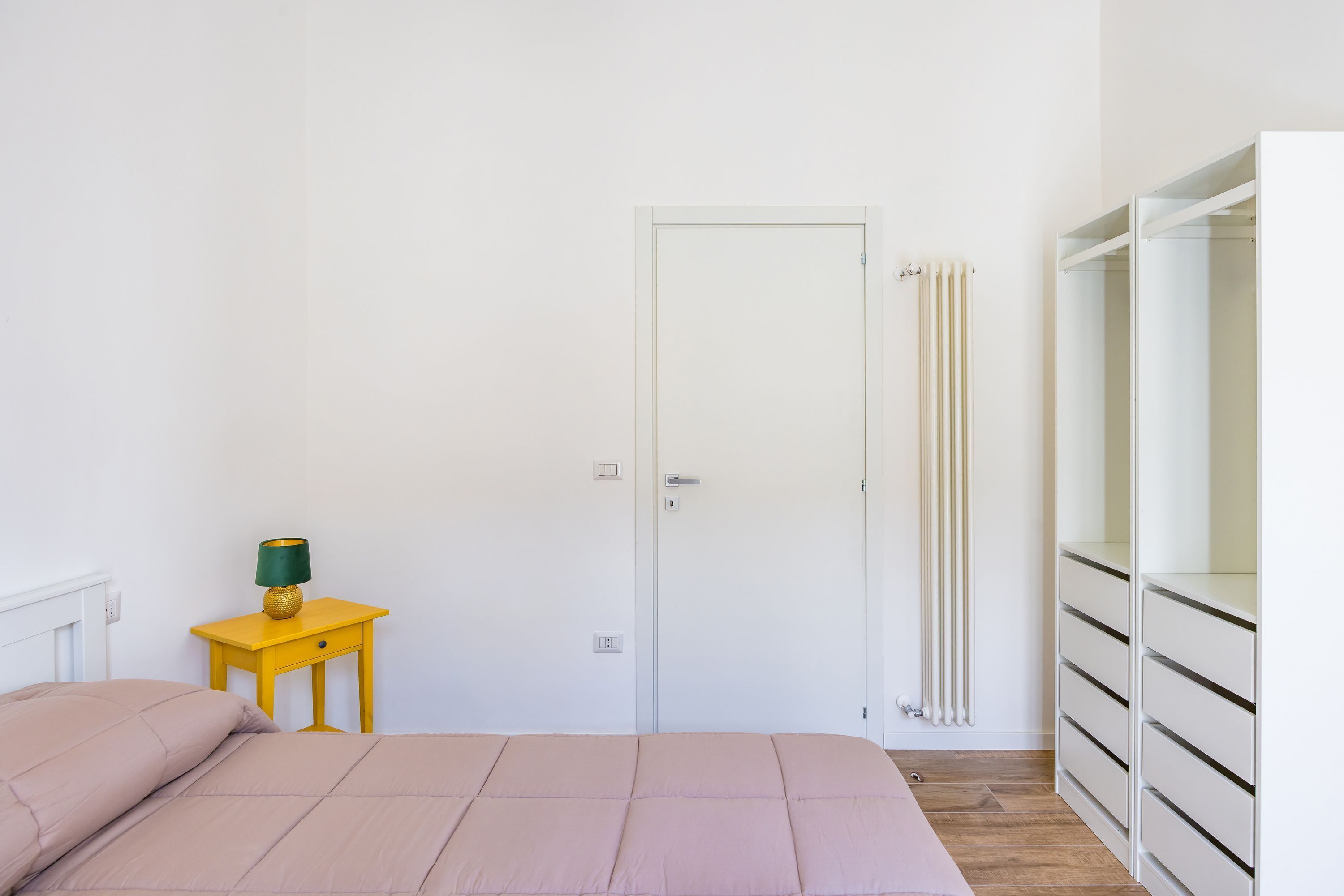 Een slaapkamer met een bed, nachtkastje met lamp, een witte deur, een radiator en een kledingkast.