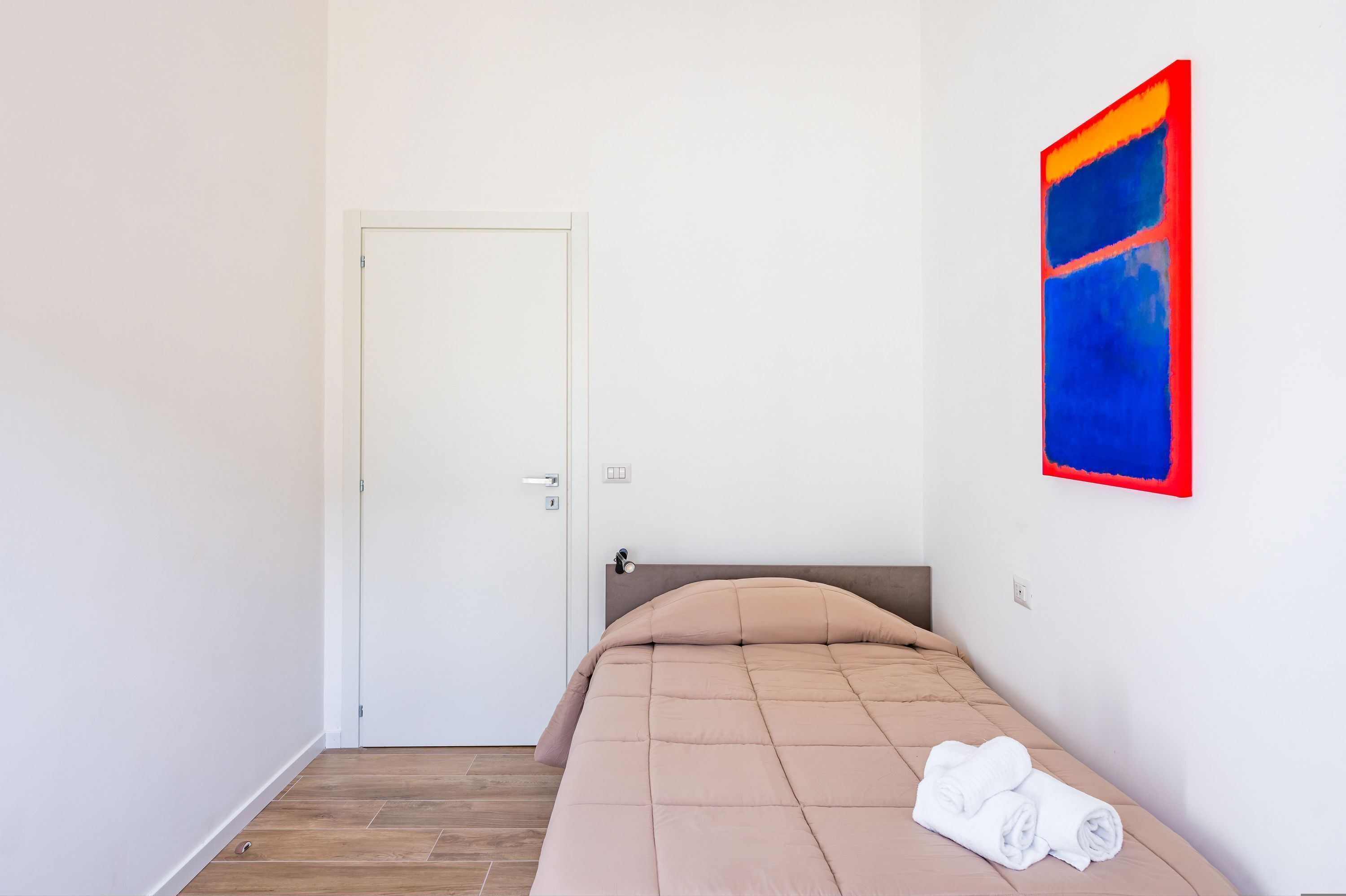 Une petite chambre à coucher, bien rangée, avec un lit simple, une porte et une œuvre d’art accrochée au mur.
