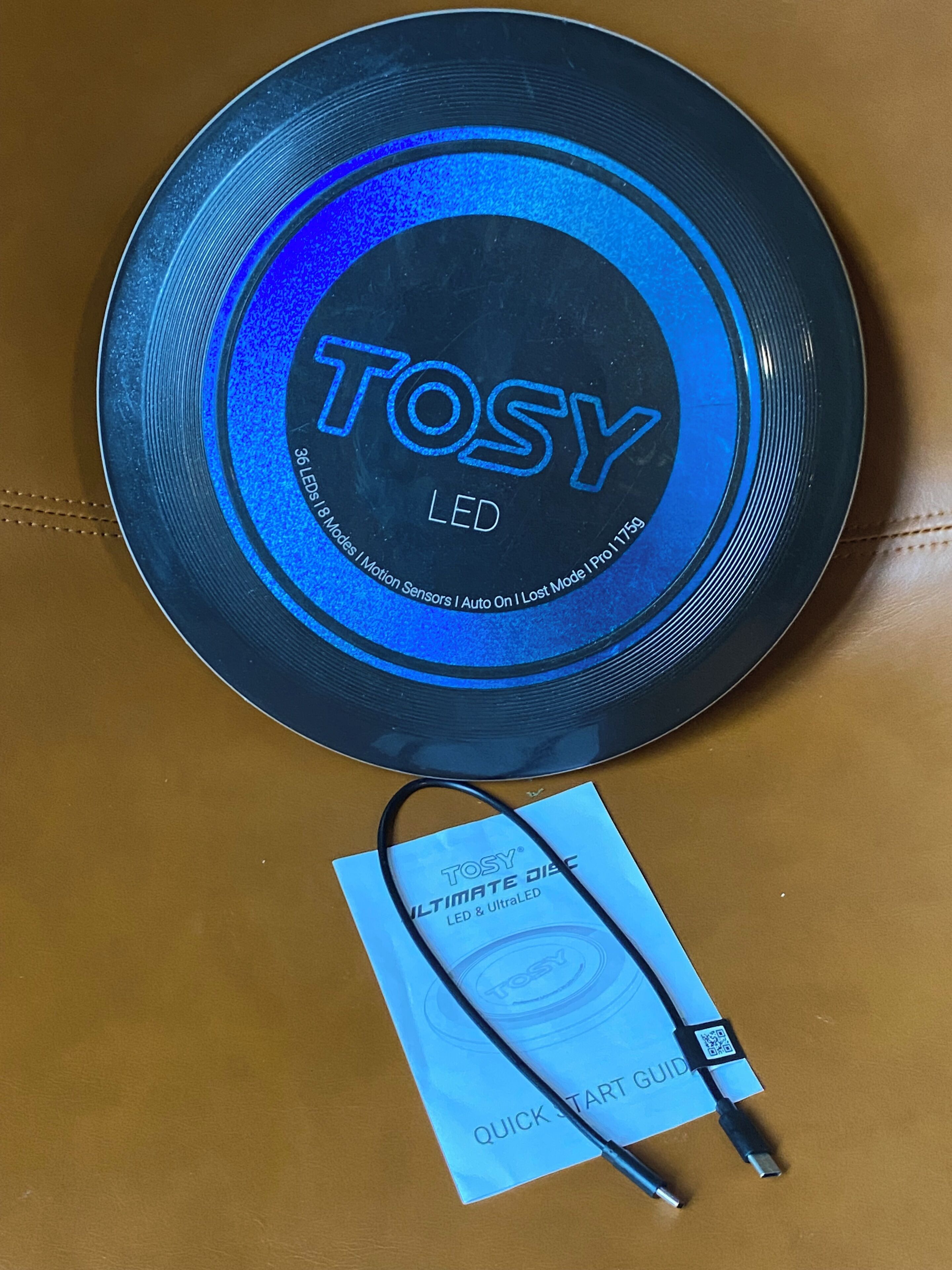 En rund genstand med teksten 'TOSY LED', et USB-kabel og en 'QUICK START GUIDE'-hæfte på en brun overflade.