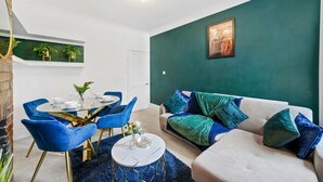 A modern living room with a round dining table, blue chairs, a beige sofa with green cushions, and a green accent wall.
