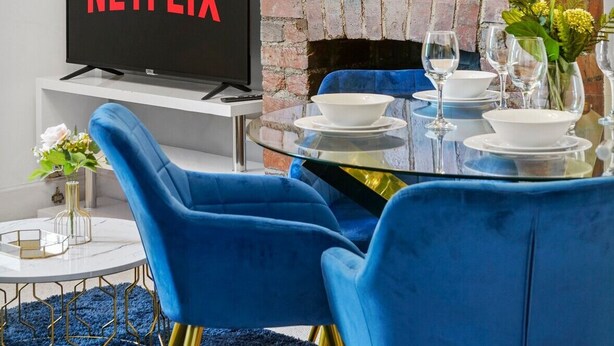 A modern living room with a brick wall, a round mirror, a flat-screen TV displaying 'NETFLIX', a dining table set with blue chairs, and a blue carpet.