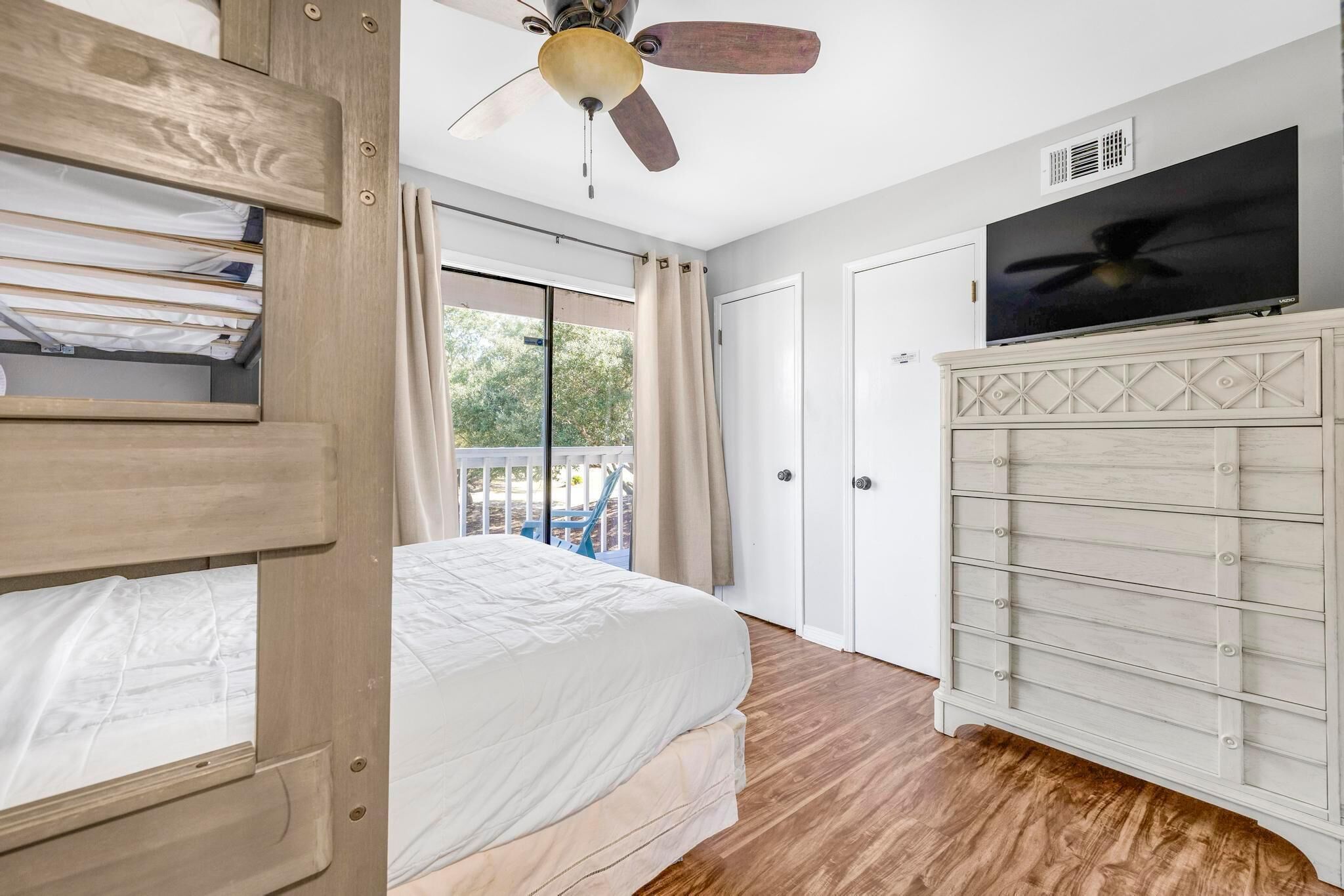 A bedroom with a bed, a ceiling fan, a TV, a wardrobe, and a sliding glass door to a balcony.