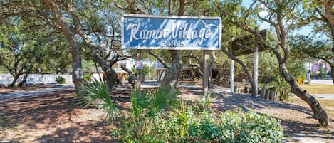 Segnaletica per 'Romer Village', con una struttura in legno rustica e vegetazione lussureggiante sullo sfondo.