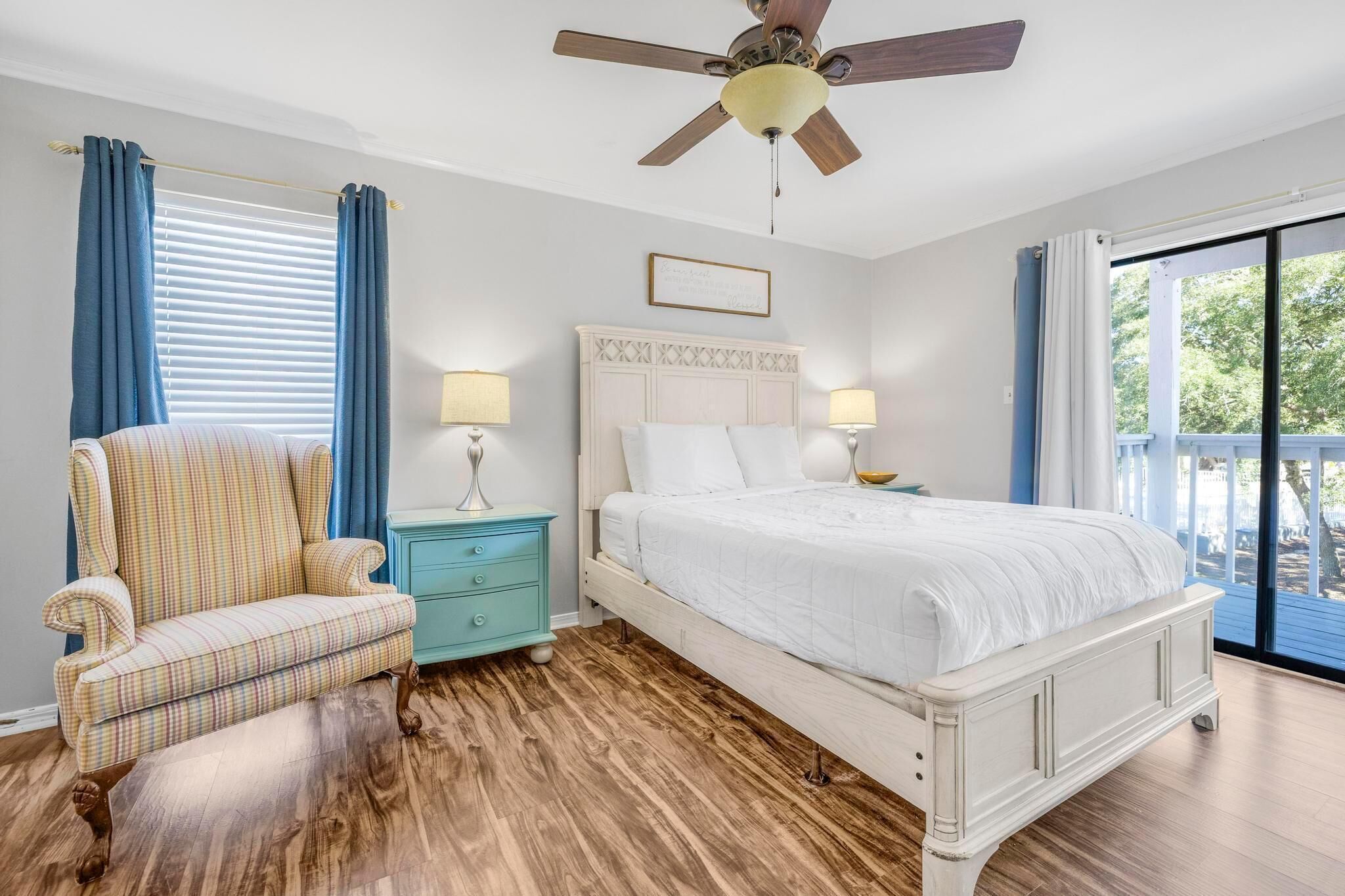 A bedroom with a bed, a chair, a nightstand, a ceiling fan, and a view of the outdoors.
