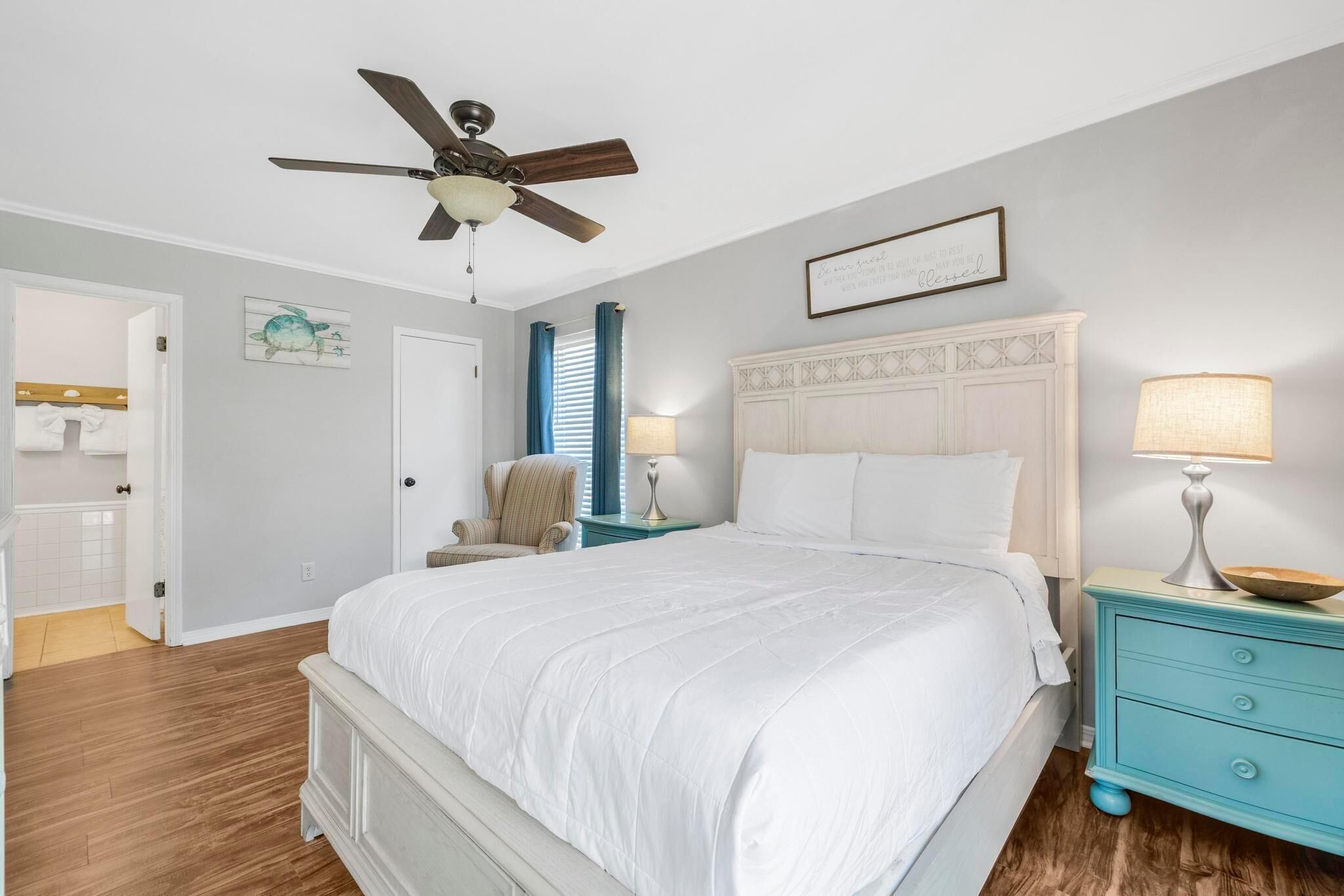 A bedroom with a bed, nightstands, a ceiling fan, a chair, and a door leading to a bathroom.