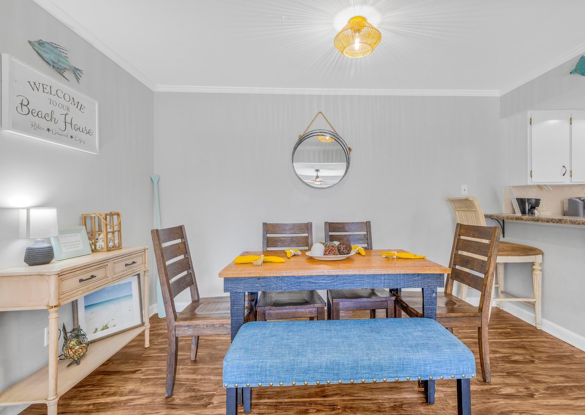 A dining area with a wooden table, four chairs, a blue bench, a wall-mounted mirror, and a ceiling light fixture.
