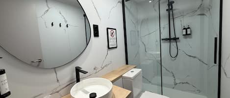 A modern bathroom with a marble wall, a round sink, a toilet, and a glass-enclosed shower.