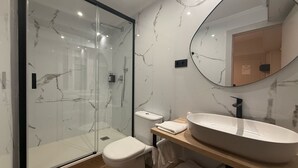 Ein modernes Badezimmer mit einer Duschkabine aus Glas, einer weißen Toilette, einem großen, ovalen Waschbecken und einem runden Spiegel an der Wand.