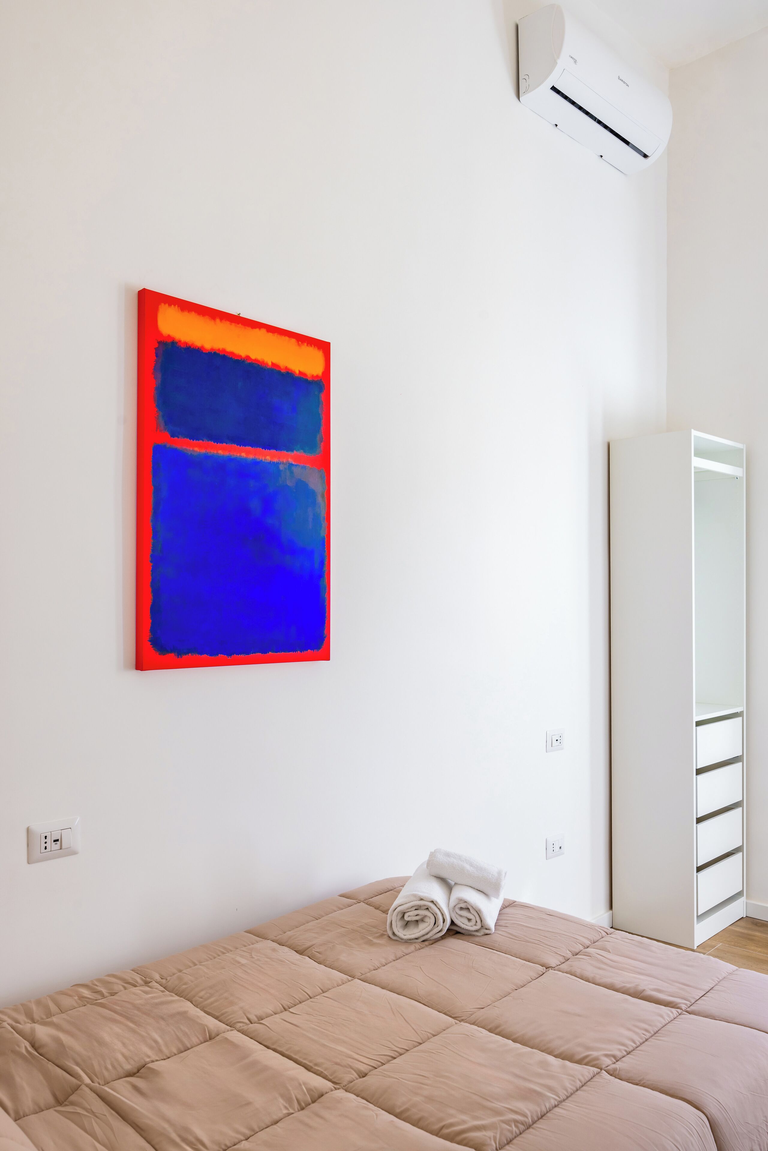 A bedroom with a bed, a wall-mounted air conditioner, a large mirror, and a red and blue abstract painting.