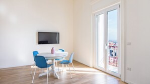 A modern living room with a white round table, blue chairs, a flat-screen TV mounted on the wall, and a large sliding glass door leading to a balcony with a city view. - Sunny Panoramic Apartment by Napoliapartments (Naples)