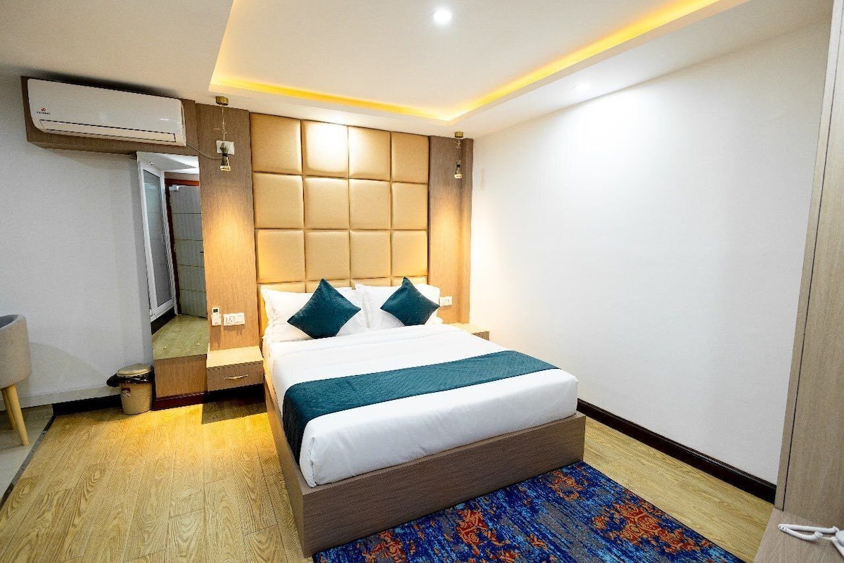 A hotel room with a large bed, a wooden headboard, a bedside table, an air conditioning unit, and a patterned rug.