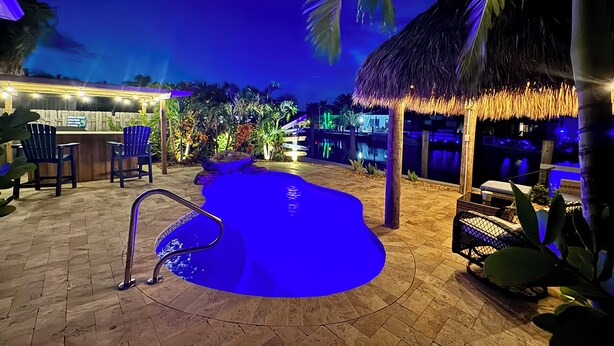Un espace piscine agrémenté d’un élément décoratif aquatique illuminé de lumière bleue, un bar avec des chaises et une structure surmontée d’un toit de chaume.