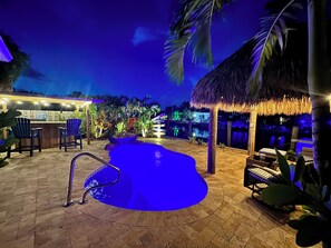 Ein Poolbereich mit beleuchtetem Wasserspiel, einer Bar mit Sitzgelegenheiten und einem strohgedeckten Gebäude.