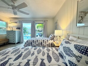 A hotel room with a bed, a nightstand, a dresser, a ceiling fan, and a view of a pool and palm trees.