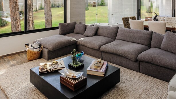 A modern living room with a sectional sofa, a coffee table, and large windows offering a view of trees and a backyard. - Casa María (Valle de Bravo)