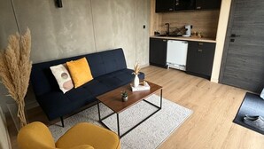 Een moderne woonkamer met een bank, salontafel en een geel fauteuil. In de achtergrond is een keuken zichtbaar met een magnetron en kastjes.