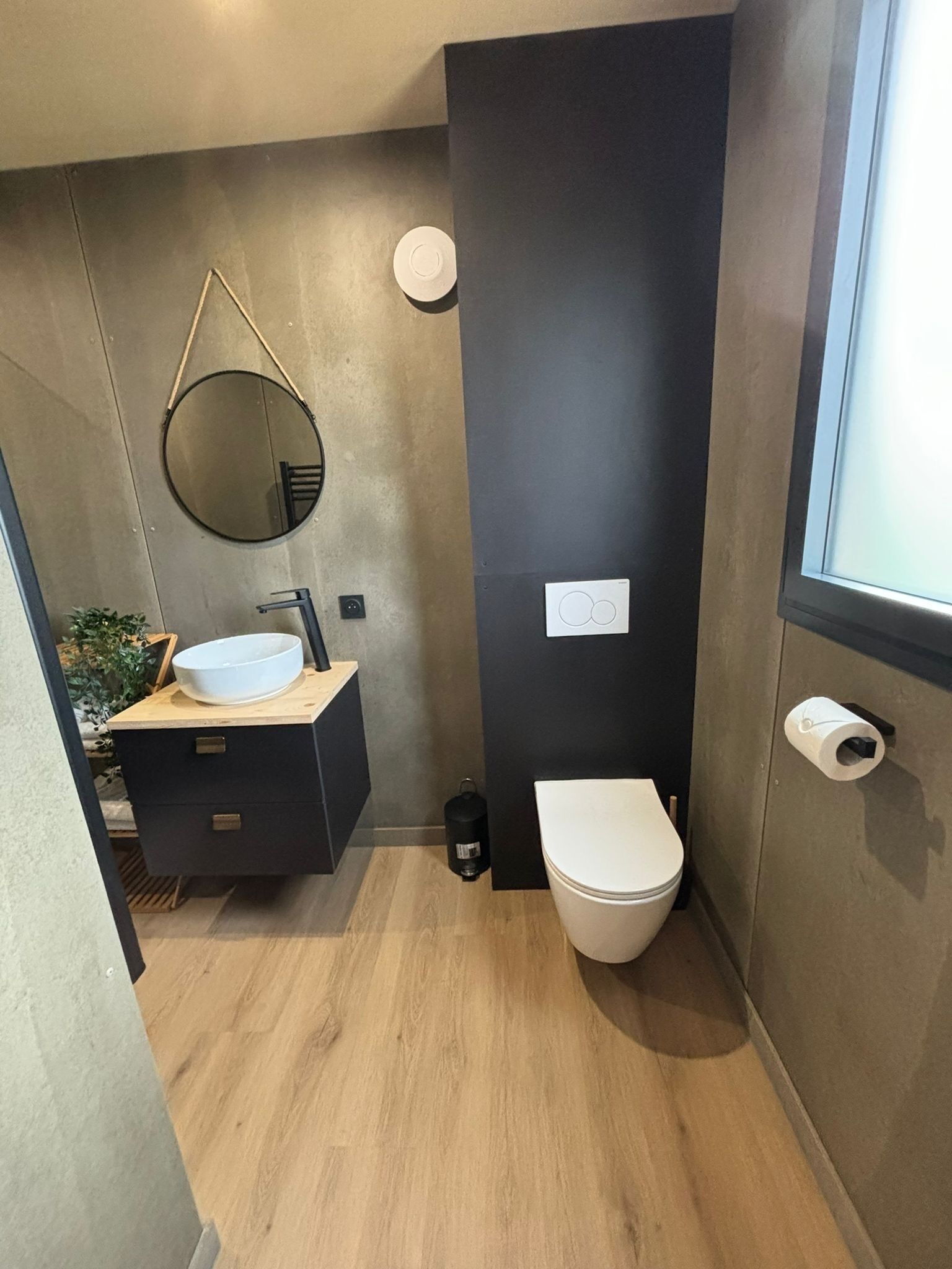 Une petite salle de bain avec un miroir rond, un lavabo blanc, des toilettes et une petite poubelle noire.