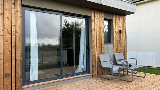 Een modern huis met een grote schuifpui, een houten terras en twee loungestoelen.