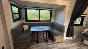 A compact RV interior with a built-in bench seating area, a small table, and two windows providing natural light and views of greenery.