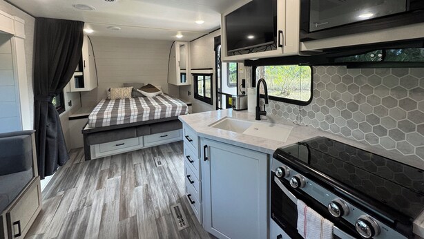 A compact RV interior with a kitchenette, a bed with a checkered blanket, and a window providing a view of the outdoors.