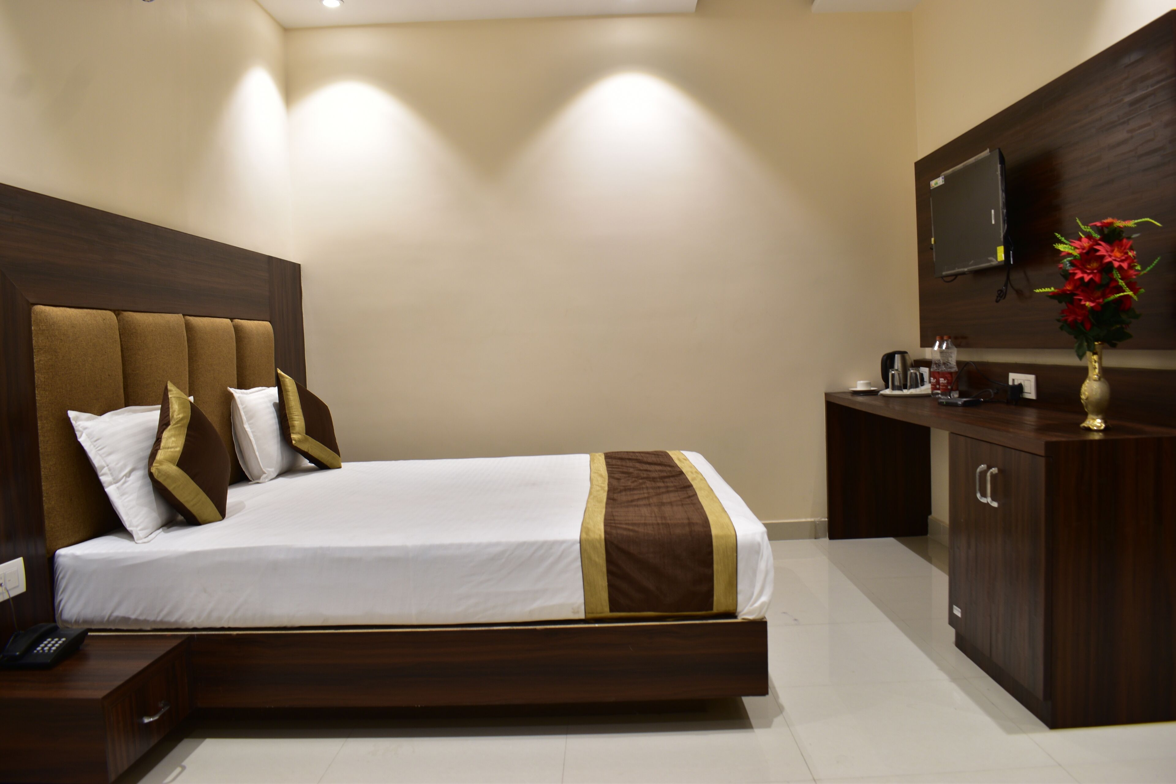 A hotel room with a large bed, a wooden headboard, a bedside table with a phone, a desk with a TV, and a cabinet.