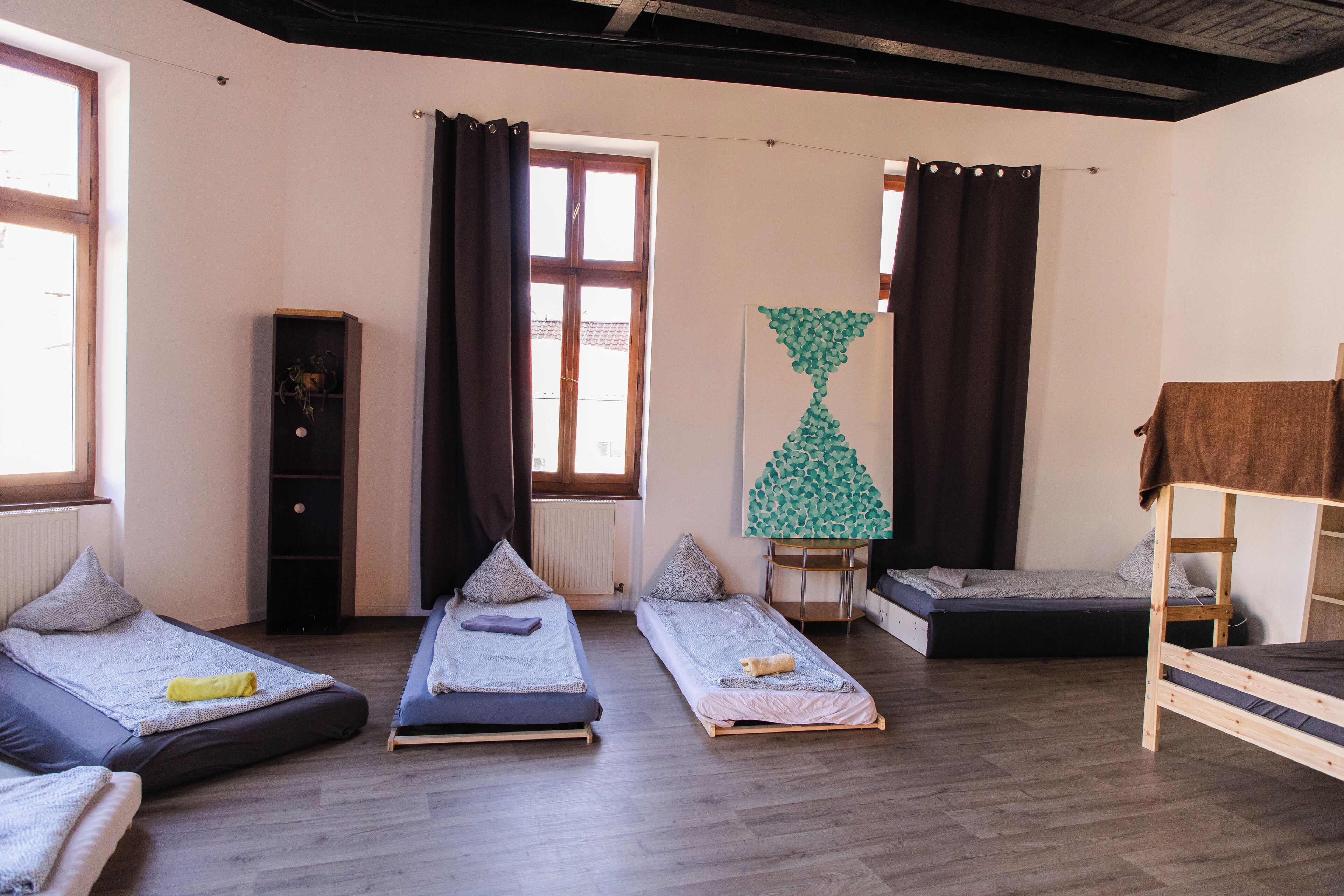 A dormitory room with three single beds, a wardrobe, a radiator, and a large window with curtains.