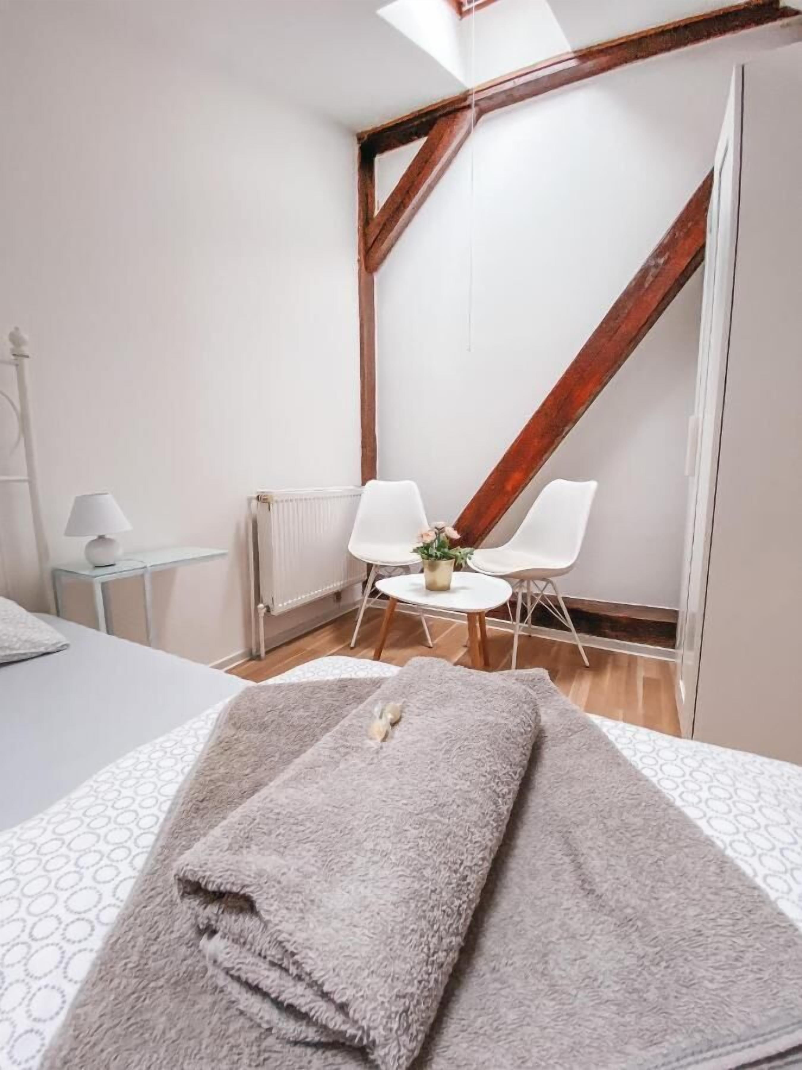 A bedroom with a bed, a small table with a lamp, a chair, and a view of a wooden staircase.