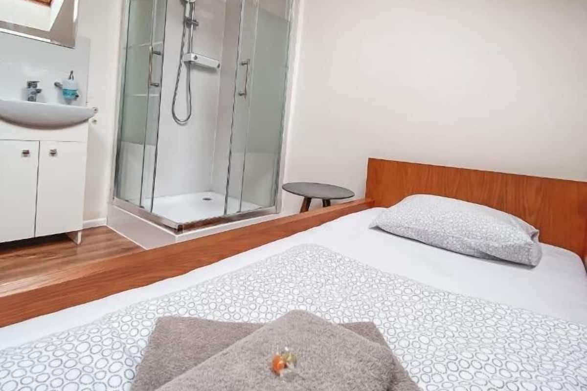 Une chambre d’hôtel comprenant un lit, une tête de lit en bois, une table de chevet, un espace douche, un lavabo et un miroir.