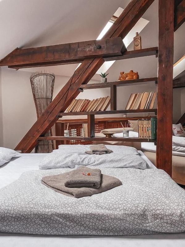 Acogedor dormitorio en ático con vigas de madera a la vista, cama alta y estantería llena de libros y objetos decorativos.