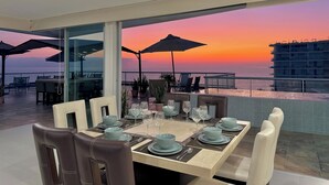 Een eetruimte met een tafel voor zes personen, met uitzicht op een zonsondergang boven de zee.