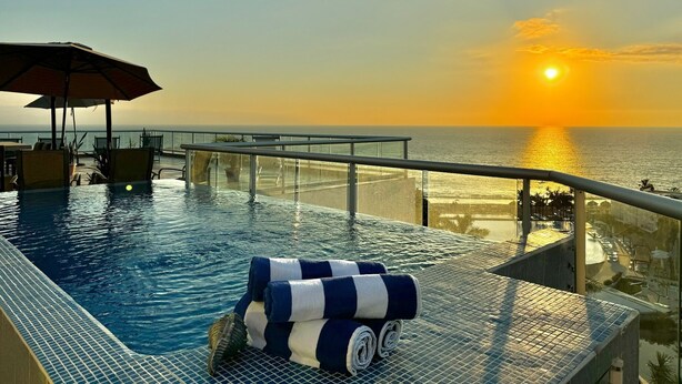 Een dakterraszwembad met een uitzicht op de ondergaande zon boven de oceaan, ligbedden en een glazen balustrade.