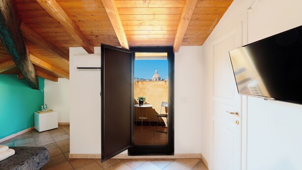 A room with a wooden ceiling, a TV, a small table with a chair, and a view of a building through an open door. - Currò Palace (Catania)