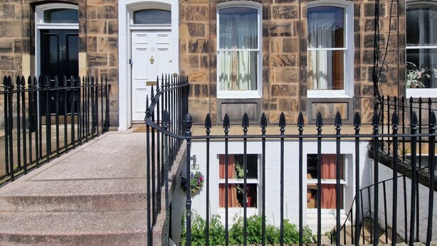 A two-story stone building with a white door and windows, a black iron fence, and steps leading to the entrance. - Gardner's Crescent City Apartment (Edinburgh)
