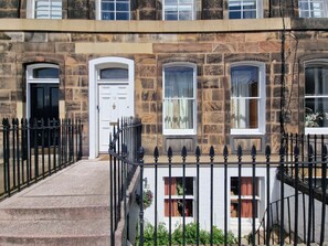 A two-story stone building with a white door and windows, a black iron fence, and steps leading to the entrance. - Gardner's Crescent City Apartment (Edinburgh)