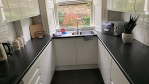 A compact kitchen with white cabinets, a black countertop, a sink, and a window with a view of greenery outside. - Gardner's Crescent City Apartment (Edinburgh)