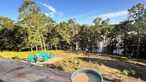 Une table en bois, sur laquelle repose une tasse de café, entourée de chaises vertes, le tout se dressant sur un fond de verdure composé d’arbres et d’un lac.
