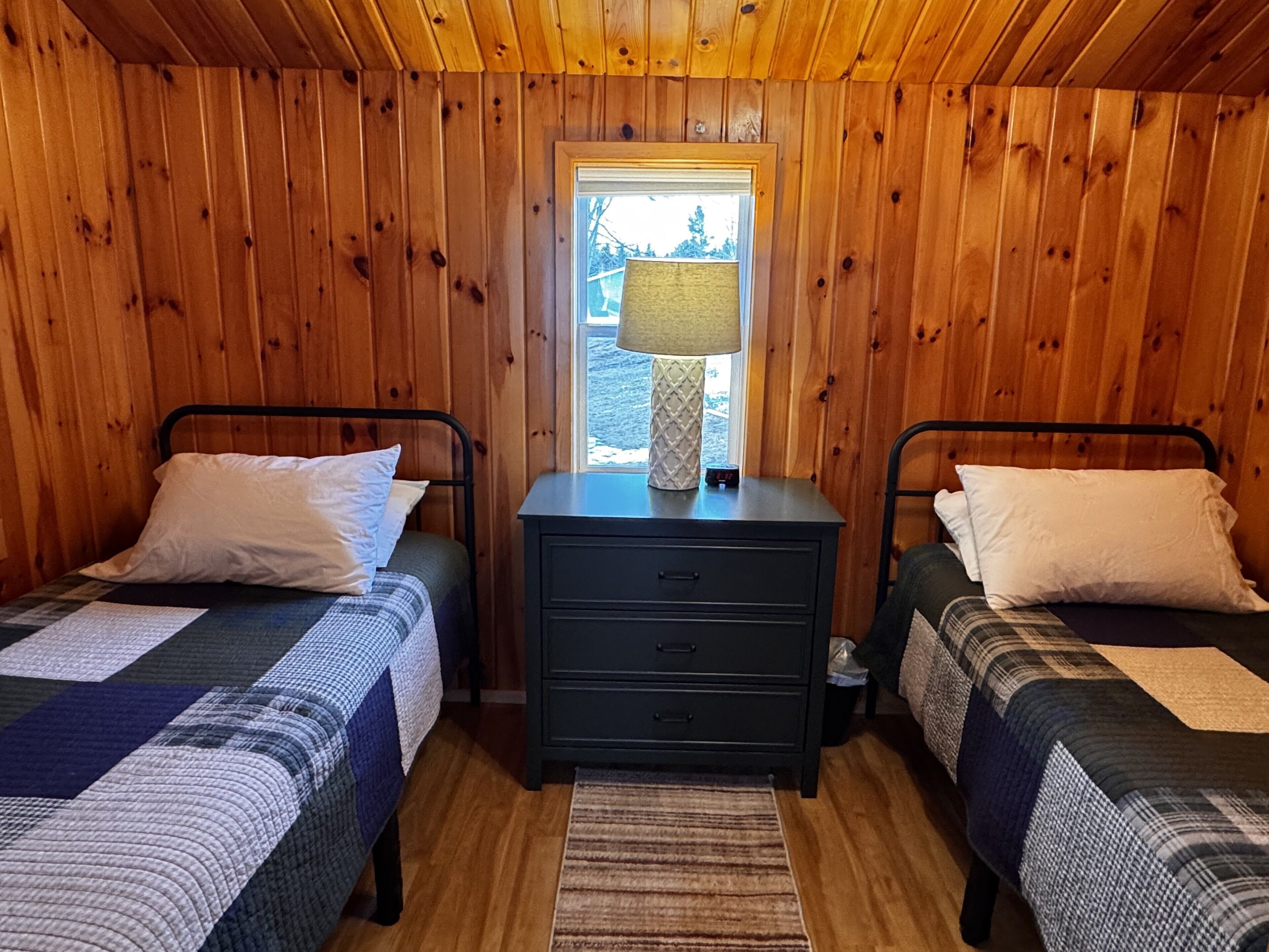 Habitación tipo cabaña con dos camas, una mesita de noche con lámpara y una ventana con vista a árboles.